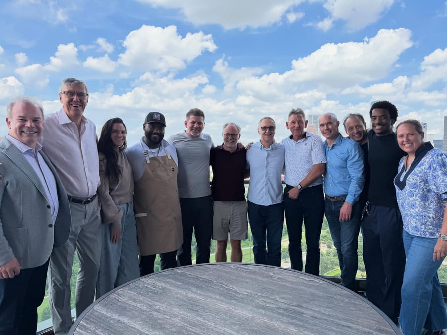 Twelve people standing arm in arm on a high terrace, with clouds and a city skyline behind them.