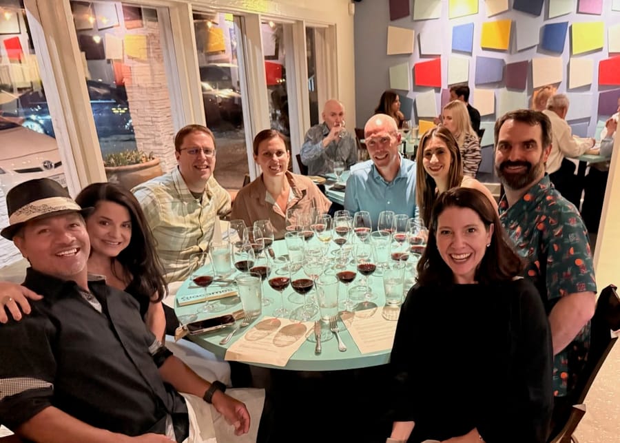 Eight people around a restaurant table lined with wine glasses, leaning in for the photo.