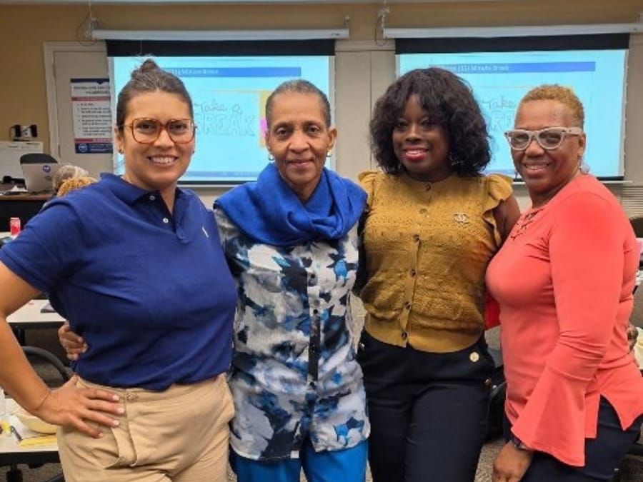 Four women standing together in a training room, with slides on the screens behind them.