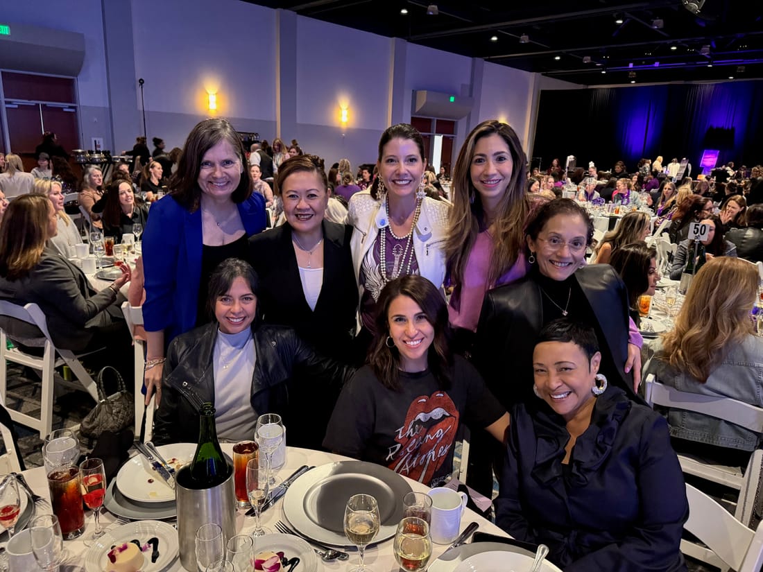 Eight women gathered at a round table at a busy luncheon, smiling at the camera.